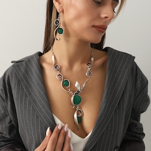 May include: A silver necklace and earring set with green gemstone details. The necklace has a large, round green stone pendant and an abstract design. The earrings have a similar design with a single green stone. The jewellery is worn with a grey blazer.