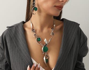 May include: A silver-toned necklace and earring set featuring green gemstone accents. The necklace has a pendant with a large, round green stone and a flowing, abstract design. The earrings have a similar design with a single green stone. The jewelry is worn with a gray blazer.