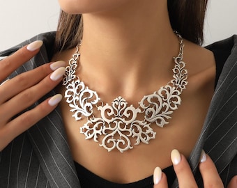 May include: A silver-toned statement necklace with an intricate, openwork floral design. The necklace features a series of interconnected, swirling patterns that create a bold, eye-catching look. The necklace is worn over a black top and a gray pinstripe blazer.