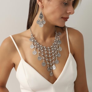 Op de afbeelding: Zilverkleurige ketting- en oorbellenset. De ketting heeft meerdere strengen kleine kralen en muntvormige hangers. De oorbellen hebben een bloemmotief aan de bovenkant met munthangers. De sieraden worden gedragen met een witte jurk.