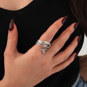 Op de afbeelding: Een zilveren ring met een uniek, abstract ontwerp. De ring is gemaakt van meerdere dunne draden die gedraaid en met elkaar verweven zijn om een vloeiende, organische vorm te creëren.