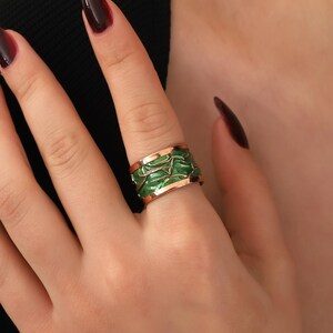 May include: A wide, gold-toned ring with a green enamel design. The ring features a textured, organic pattern in a vibrant green colour, set against the warm gold metal. The ring is worn on a finger.