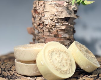 May include: Three white round bars of soap with a textured surface, sitting on a woven brown wicker mat. The soap is wrapped in clear plastic.