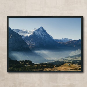 Puede incluir: Una fotografía enmarcada de una cordillera con picos cubiertos de nieve y un valle lleno de niebla. Las montañas son de color azul oscuro y la niebla es de color azul claro. El cielo es de color azul claro.