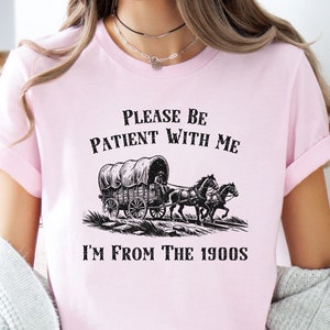 May include: Pink t-shirt with a black graphic of a covered wagon pulled by two horses. The text "Please be patient with me I'm from the 1900s" is printed on the shirt.