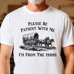 May include: White t-shirt with a black and white illustration of a covered wagon pulled by two horses. The text "Please be patient with me I'm from the 1900s" is printed on the shirt.