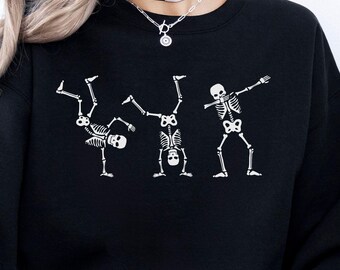 May include: A black long-sleeve sweatshirt featuring a white graphic print of three skeletons in different poses. One skeleton is performing a one-handed handstand, another is in a handstand, and the third skeleton is performing a dabbing motion.