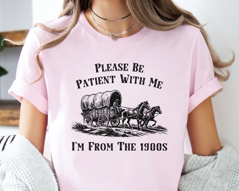 May include: Pink t-shirt with a black graphic of a covered wagon pulled by two horses. The text "Please be patient with me I'm from the 1900s" is printed on the shirt.