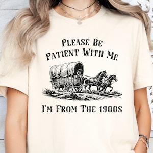 May include: A white t-shirt with black text that reads "Please be patient with me I'm from the 1900s". The shirt features a black and white illustration of a covered wagon pulled by two horses.