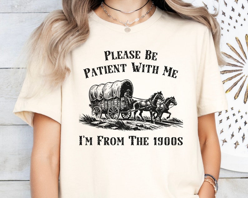 May include: A white t-shirt with black text that reads "Please be patient with me I'm from the 1900s". The shirt features a black and white illustration of a covered wagon pulled by two horses.