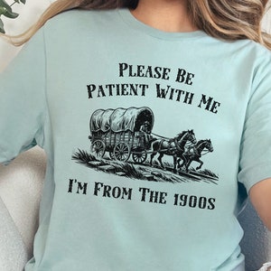 May include: A light blue t-shirt with a black graphic of a covered wagon pulled by two horses. The text "Please be patient with me I'm from the 1900s" is printed on the shirt.