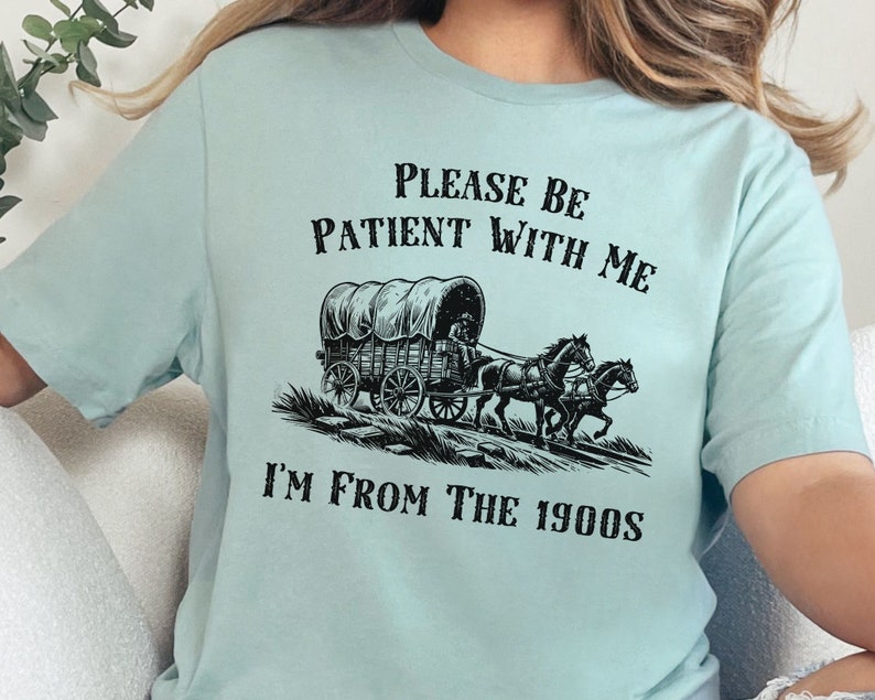 May include: A light blue t-shirt with a black graphic of a covered wagon pulled by two horses. The text "Please be patient with me I'm from the 1900s" is printed on the shirt.