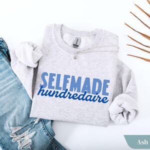 May include: A light gray sweatshirt with the text "SELFMADE hundredaire" in blue. The sweatshirt is paired with blue jeans and a silver necklace. The word "Ash" is visible in the bottom right corner.