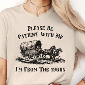 May include: A beige t-shirt with black text that reads "Please be patient with me I'm from the 1900s". The text is above a black and white illustration of a covered wagon pulled by two horses.
