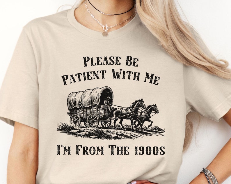 May include: A beige t-shirt with black text that reads "Please be patient with me I'm from the 1900s". The text is above a black and white illustration of a covered wagon pulled by two horses.