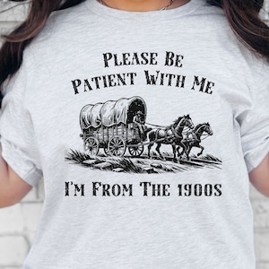 May include: A grey t-shirt with a black graphic of a covered wagon pulled by two horses. The text "Please be patient with me I'm from the 1900s" is printed on the shirt.