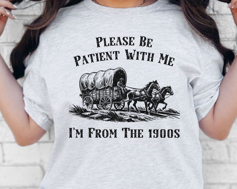 May include: A grey t-shirt with a black graphic of a covered wagon pulled by two horses. The text "Please be patient with me I'm from the 1900s" is printed on the shirt.