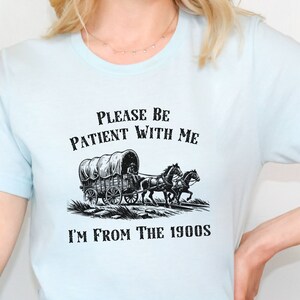 May include: A light blue t-shirt with a black and white illustration of a covered wagon pulled by two horses. The text "Please be patient with me I'm from the 1900s" is printed on the shirt.