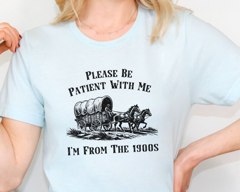 May include: A light blue t-shirt with a black and white illustration of a covered wagon pulled by two horses. The text "Please be patient with me I'm from the 1900s" is printed on the shirt.