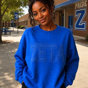 May include: A vibrant royal blue crewneck sweatshirt featuring the word ZETA embossed in a matching tonal font across the chest. The garment has long sleeves with ribbed cuffs and a ribbed neckline, providing a clean and structured appearance.