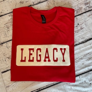 Peut inclure: T-shirt rouge avec un rectangle blanc sur lequel est inscrit "LEGACY" en caractères blancs gras.