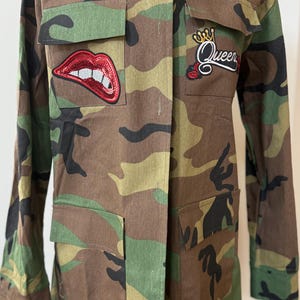Può includere: Giacca con stampa mimetica con maniche lunghe e tasche multiple. Presenta una toppa di labbra rosse con paillettes e una toppa "Queen" con un design a corona e rosa. La giacca è esposta su un manichino nero.