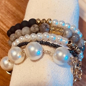 Puede incluir: Un conjunto de pulseras con cuentas negras, grises y blancas, incluyendo grandes perlas blancas y detalles dorados.