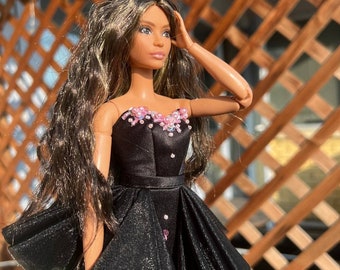 May include: A doll with long brown hair wearing a black dress with a flared skirt. The dress is embellished with pink beads around the neckline and hem. The doll is posed with one hand on its head.