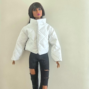 May include: A doll dressed in a white quilted jacket with a high collar and black ripped jeans. The jacket features a diamond pattern and a zip. The doll has dark hair and is wearing makeup.