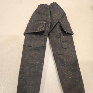 May include: Dark grey cargo trousers with two square pockets on each leg. The trousers have an elasticated waistband and are made of a textured fabric. The trousers are designed for a doll.