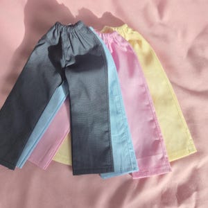 May include: Four pairs of doll trousers in various colours: grey, light blue, pink, and yellow. The trousers have an elasticated waistband and a straight leg design. They appear to be made from a smooth, possibly satin-like fabric.
