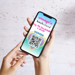 May include: A smartphone displays a screen with the text "amaya's bachisland buy the bombshell a drink!" and a Venmo QR code. The screen has a gradient background with a heart-shaped design. The phone is held in two hands.