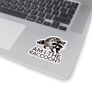 May include: A die-cut sticker featuring a detailed illustration of a raccoon with grey, black, and white fur. Below the raccoon, the text AM I THE RACCOON? is printed in bold, black, sans-serif capital letters with a subtle pinkish shadow effect.
