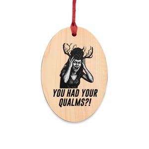 May include: An oval-shaped light wood ornament hanging from a red ribbon. The center features a black ink illustration of a person with antlers on their head, looking distressed with hands on their face. Below the image is the text YOU HAD YOUR QUALMS?!