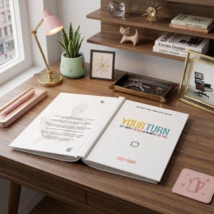 May include: An open hardcover book titled Your Turn by Lucy Fours with white pages and black, yellow, teal, and red text. The book rests on a wooden desk alongside a pink lamp, a green potted plant, a tray with keys, and a pink coaster.