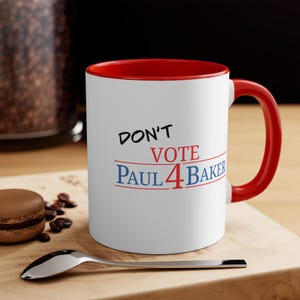 Op de afbeelding: Een witte keramische mok met een rode binnenkant en handvat. De voorzijde bevat de zwarte tekst DON'T, rode tekst VOTE en blauwe tekst PAUL 4 BAKER, met rode en blauwe horizontale lijnen onder de woorden VOTE en PAUL 4 BAKER.