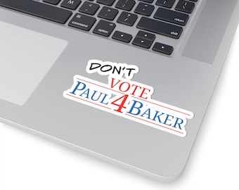 May include: A rectangular sticker with a white border featuring the black text DON'T, red text VOTE, and blue text PAUL 4 BAKER. The sticker is placed on the silver surface of a laptop keyboard, displaying a political-style graphic design.
