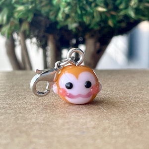 May include: A charm featuring a monkey face with orange, pink, and white details. The charm has a silver-colored clasp. The monkey's face has black eyes and a pink mouth. The charm is likely for a bracelet or necklace.