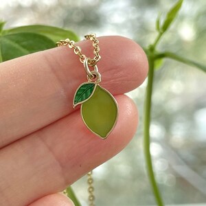 May include: A gold chain necklace with a green enamel lemon charm. The lemon has a small green leaf attached to the top.