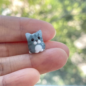 Puede incluir: Una peque&ntilde;a figurita de vidrio de un gato gris y blanco con ojos negros. El gato est&aacute; sentado con las patas metidas.