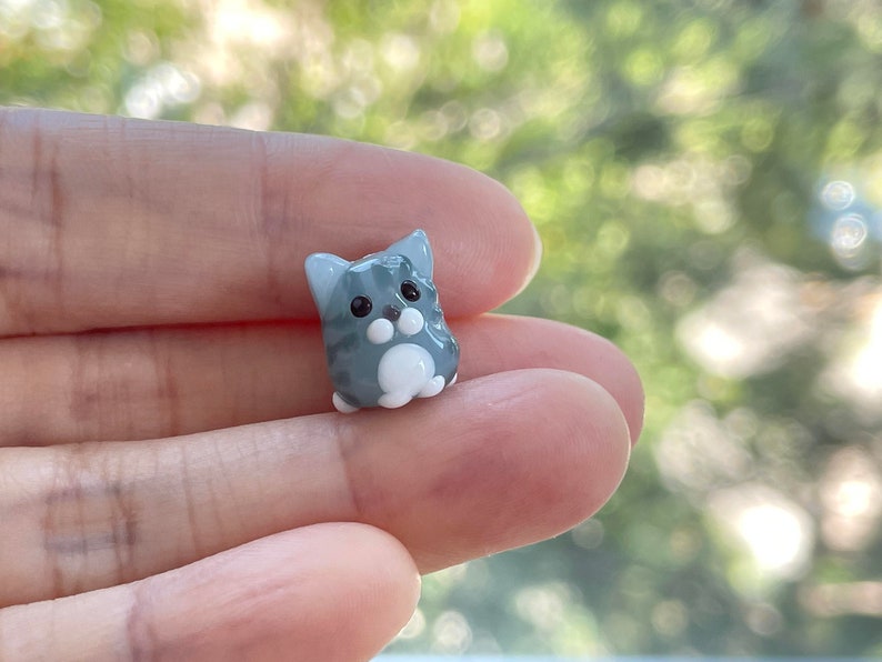 Puede incluir: Una peque&ntilde;a figurita de vidrio de un gato gris y blanco con ojos negros. El gato est&aacute; sentado con las patas metidas.