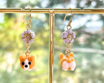 以下が含まれることがあります： 紫色の花チャームに金色のアクセントが施された、茶色と白のコーギーチャームが付いた、ぶら下がりのイヤリングのペアです。