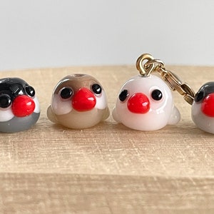 May include: Four small glass bird figurines with black eyes and red beaks. Three are sitting on a wooden surface, while the fourth is hanging from a gold chain.