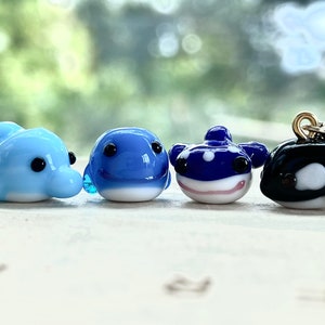 May include: Four small glass animal figurines, a blue dolphin, a blue whale, a blue and white spotted pufferfish, and a black and white orca. The figurines are all facing forward and are arranged in a row.