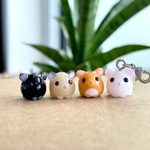 May include: Four small glass hamster figurines with different coloured bodies. The hamsters are black, beige, orange, and white. The white hamster is attached to a silver charm.