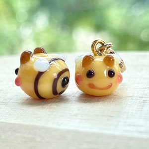 May include: Two small, yellow bee-shaped charms with brown stripes and details. One has a gold-coloured loop for hanging. The bees have black eyes, pink cheeks, and white accents. The background is a blurred green.