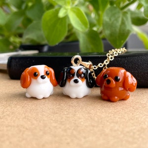 May include: Three glass dog charms, one on a gold chain. The charms are white, black, and brown, and are shaped like Cavalier King Charles Spaniels.