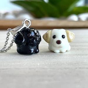 May include: A black dog-shaped glass pendant attached to a silver-toned metal chain, positioned next to a separate white and tan dog-shaped glass bead. Both items feature small black eyes and a brown nose, crafted with a smooth, glossy finish.