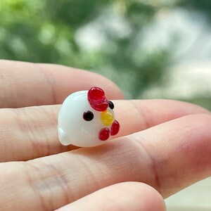 May include: A small white glass figurine of a chicken with red and yellow accents. The chicken is sitting on a hand.