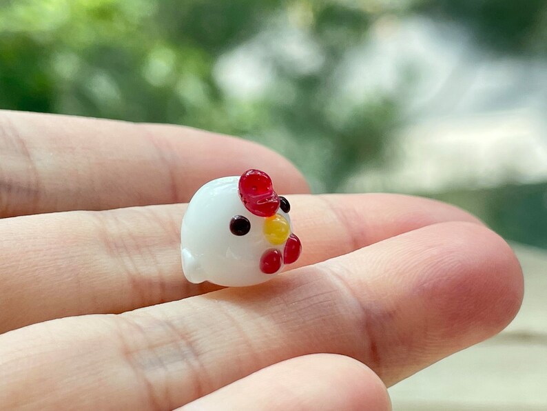 May include: A small white glass figurine of a chicken with red and yellow accents. The chicken is sitting on a hand.